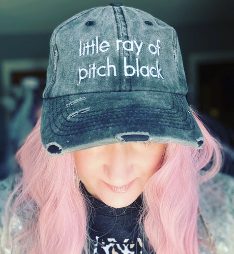 Person wearing a distressed denim cap with 'little ray of pitch black' text, pink hair, blurred background