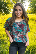 woman in a turquoise and brown western print short sleeve top with relaxed fit paired with black jeans standing outside in field