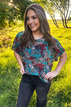 woman in a turquoise and brown western print short sleeve top with relaxed fit paired with black jeans standing outside in field