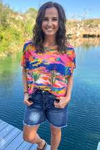 Woman in bright colored blue pink yellow green shirt paired with denim shorts standing on a porch