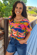 Woman in bright colored blue pink yellow green shirt paired with denim shorts standing on a porch