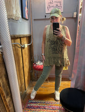 Girl in a yellowish tank top that says pretty is as pretty does taking a selfie in front of mirror wearing a hat and green pants and white sneakers