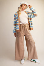 A woman models a casual outfit featuring a blue, brown, and beige striped cardigan, wide-leg tan pants, a white t-shirt, a white bucket hat, and white sneakers with green accents, set against a plain beige studio background.