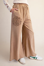 A person wearing beige wide-leg drawstring pants with a large crocheted floral detail on the upper left leg, paired with white sneakers against a plain beige studio background.