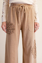 A close-up of a person wearing beige wide-leg drawstring pants with decorative crochet floral patches on the pockets and lower legs, paired with a cream long-sleeve top against a light neutral background.