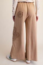A person shown from the back wearing wide-leg tan pants with embroidered floral details on the back pockets, paired with a cream top and white sneakers.