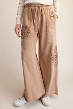 A person wearing wide-leg tan pants with embroidered floral details on the lower legs, a cream drawstring waist, and side pockets, paired with white sneakers against a plain beige studio background.