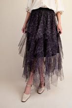 A model wearing a dark purple and black leopard-print tulle midi skirt with a layered ruffle design, paired with a cream blouse and beige heels, set against a plain beige studio background.
