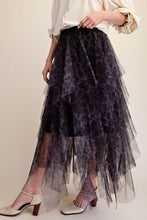 A model wearing a long, tiered tulle skirt in a dark purple and black leopard print, paired with a cream blouse and white heeled sandals, set against a plain beige background.
