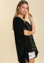 Woman wearing a black embroidered top with colorful designs on a beige background