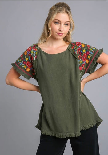 Woman wearing an olive green top with colorful embroidered sleeves on a gray background