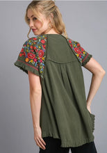 Woman wearing an olive green top with colorful floral embroidery on a gray background