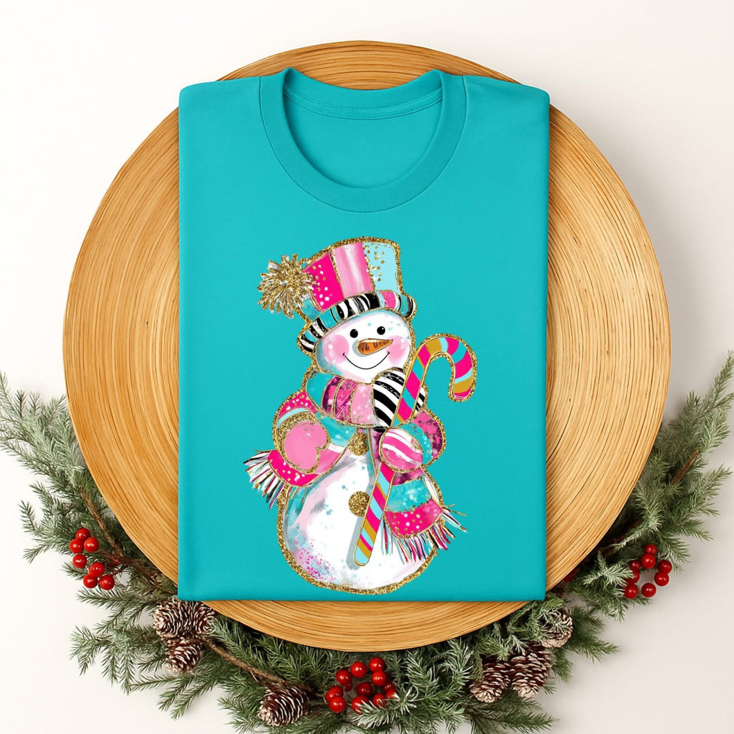 Turquoise t-shirt with a colorful snowman design on a wooden plate surrounded by Christmas decorations.