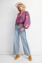 Woman wearing a purple floral blouse, light blue jeans, and a white hat on a white background