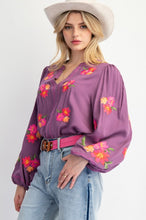 Woman wearing a purple blouse with floral embroidery, light blue jeans, and a beige hat on a white background