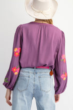 Purple blouse with floral embroidery on long sleeves worn by a person, wearing a white hat and light blue jeans.