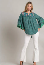 Woman wearing a green patterned blouse with floral embroidery and white pants on a plain background