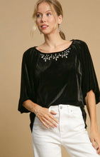 Woman wearing a black velvet top with embellishments and white pants against a beige background
