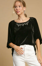 Woman wearing a black velvet top with embellishments and white pants on a beige background