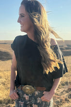 Woman standing in a field wearing a black top and patterned pants with a large belt buckle.