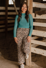 Woman wearing a teal top and leopard print pants standing in a rustic indoor setting.