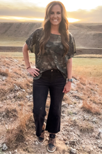 Woman standing in a field wearing a camouflage shirt and black pants, with a sunset in the background.