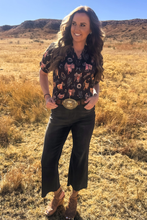 Woman standing in a field wearing a patterned shirt and dark pants with a belt buckle.