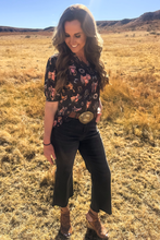 Woman standing in a field wearing a floral shirt and black pants.