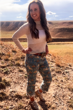 Woman standing in a desert landscape wearing a white top and patterned pants.