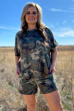 Woman wearing a camouflage outfit standing in an open field with blue sky.