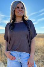 Woman wearing a brown t-shirt and light blue jeans standing in a field with a blue sky.