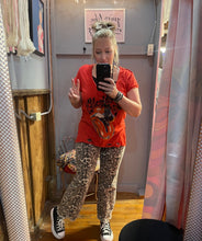 Person taking a mirror selfie wearing an orange t-shirt and leopard print pants in a store.