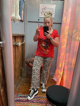Person taking a mirror selfie wearing a red shirt and patterned pants in a room with colorful curtains.