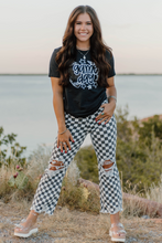 Woman wearing a black t-shirt with graphic design and checkered pants standing outdoors near water.